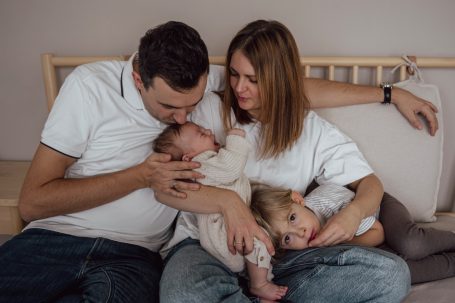 Familie sitzt auf einem Sofa, Eltern küssen das Baby, das Kleinkind kuschelt.