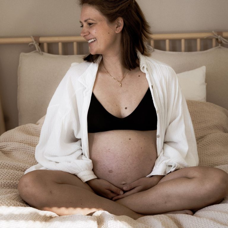 Schwangerschaftsfotografie Schwangere Frau sitzt lächelnd auf einem Bett, umgeben von weichen Kissen.