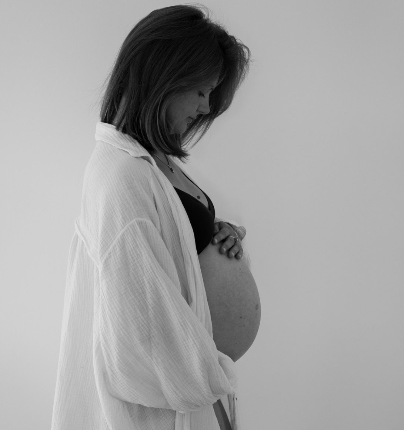 Newbornfotografie Schwangere Frau in einem weißen Oberteil, die sanft ihren Bauch hält.