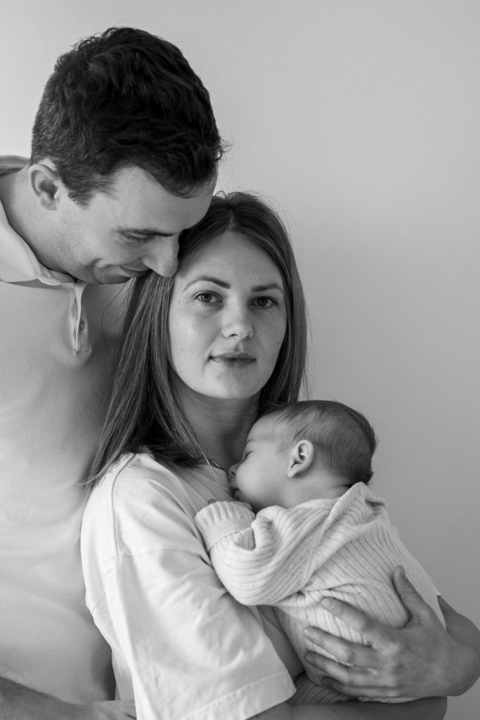 Newbornfotografie Ein Vater hält seine Partnerin und ein Baby in einer liebevollen Umarmung.