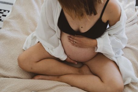 Schwangere Frau in Unterwäsche hält ihren Bauch, sitzt auf einem beigen Teppich.