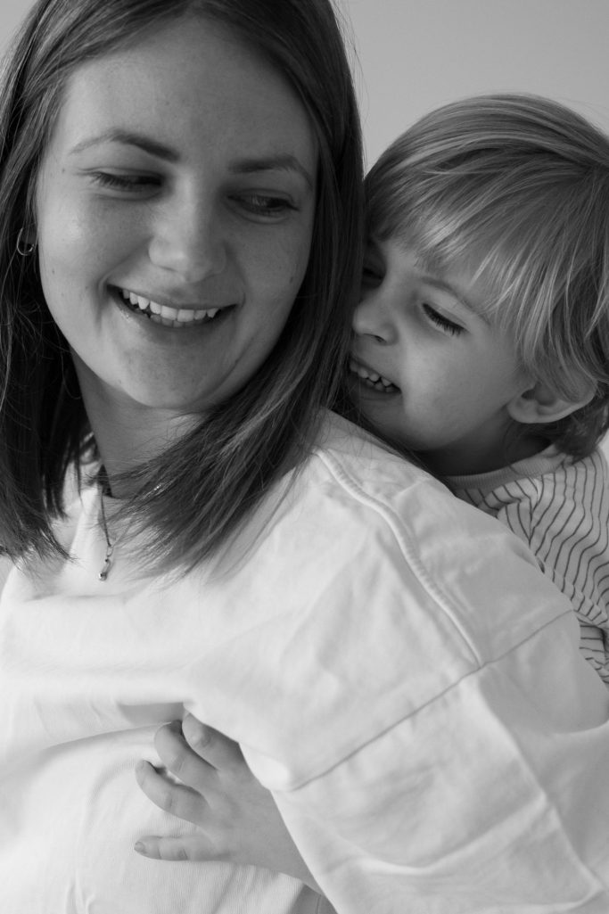Familienfotografie Frau mit langen Haaren lächelt, während ein kleines Kind ihr von hinten umarmt.